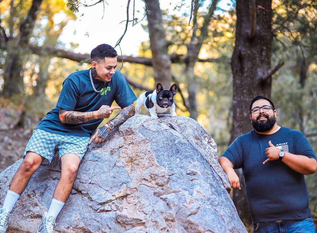 Two men smiling with a French Bulldog named Moo on a large rock in the forest.
